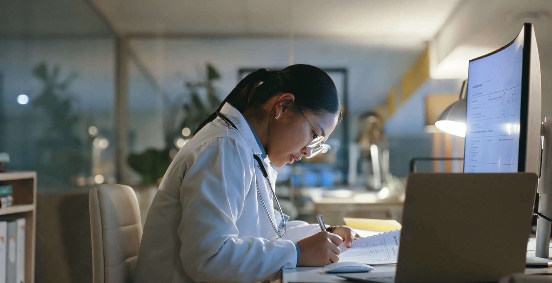 Doctor in white coat with stethoscope and glasses writing notes at a desk beside a lit computer monitor.