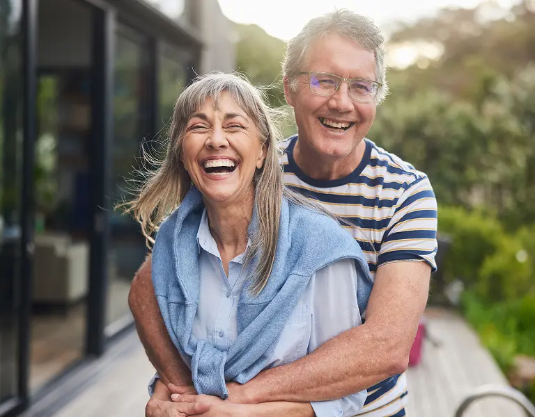 Older couple laughing on a deck, man in striped shirt hugging woman with blue sweater draped over shoulders.