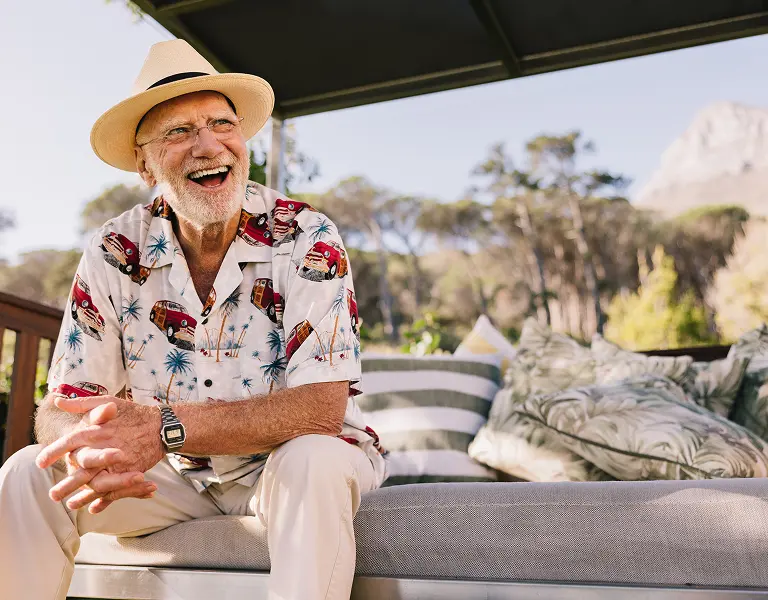 Elderly man wearing a straw hat and patterned shirt laughing while seated on an outdoor sofa with cushions and trees behind
