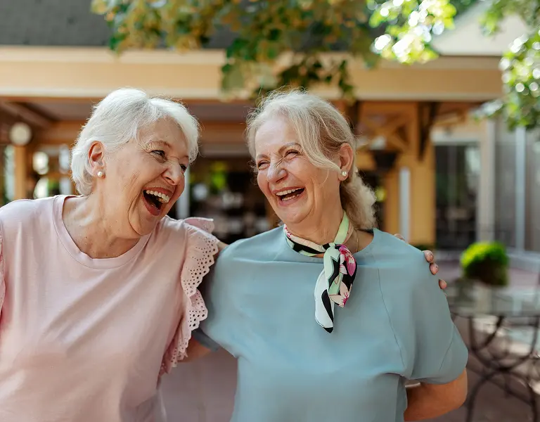 Two elderly women arm-in-arm laughing outdoors, wearing pastel tops and one with a patterned neck scarf.