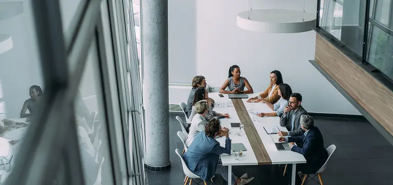 Eight colleagues gather around a long white table in a glass-walled modern boardroom, discussing documents and laptops.
