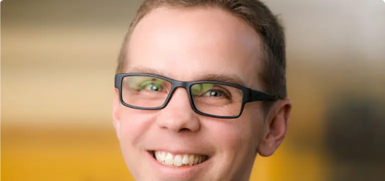 Smiling young man with short brown hair and black rectangular glasses against a soft, warm-toned blurred background.