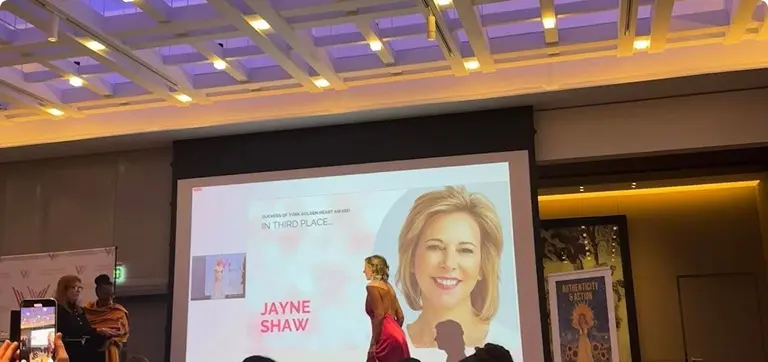 Woman in a red dress on stage receiving an award in front of a large projected portrait labeled "Jayne Shaw", audience member taking a photo