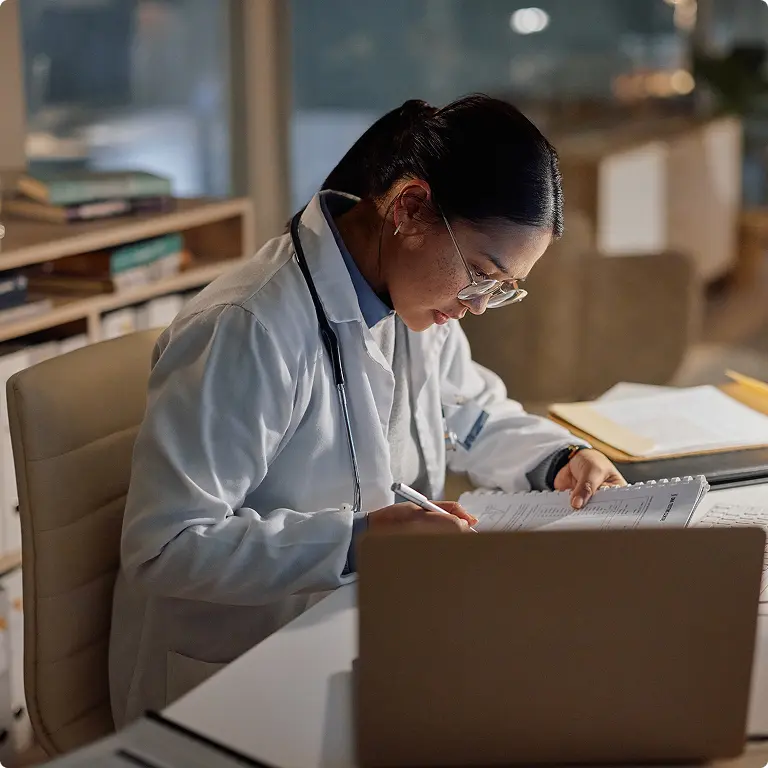 Doctor in white coat with stethoscope writing notes at a desk beside a laptop in a softly lit office.